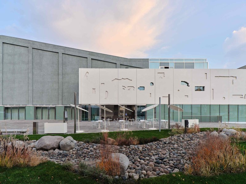 Cantilevered facade with organic cutouts above a glazed base, adjacent to a landscaped rock garden at dusk