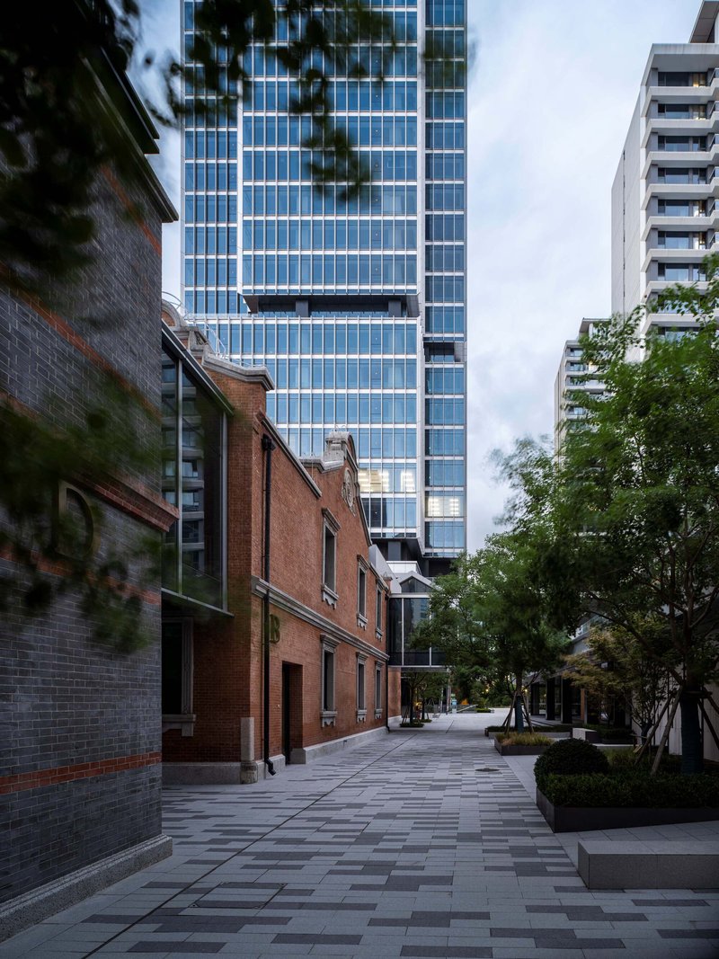 Paved pedestrian plaza between the preserved brick buildings and the glass tower at twilight