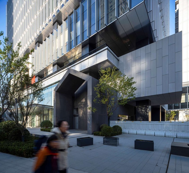 Ground-level plaza with black benches and planted beds as a person walks beneath the cantilevered volume
