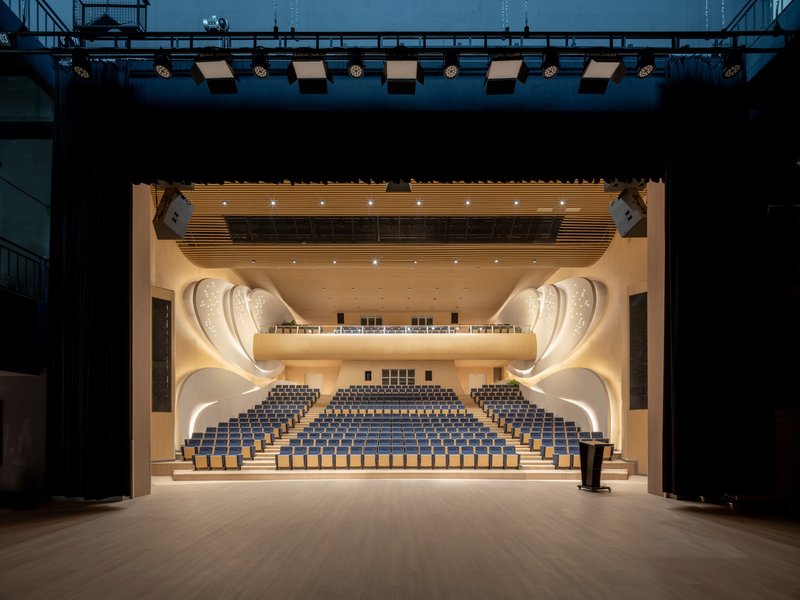 View from the stage into the auditorium showing curved balconies, timber acoustic ceiling, and rows of blue seats