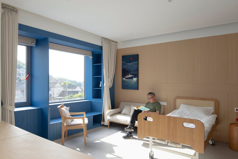 Patient room with timber wall panels, blue window alcove, and a person reading beside the bed