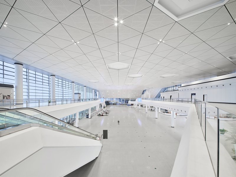 Interior departure hall with perforated white ceiling panels and glass curtain wall overlooking the tarmac