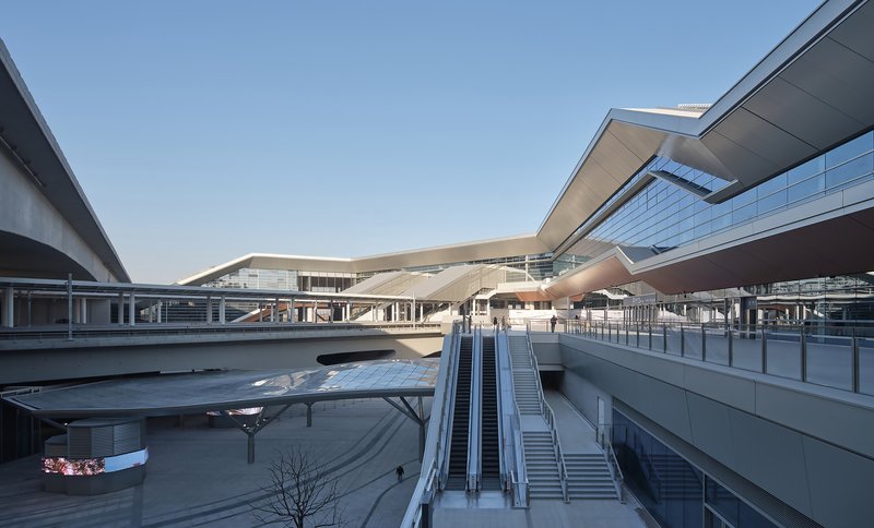 Multilevel transit plaza with escalators leading between concrete platforms under a clear sky