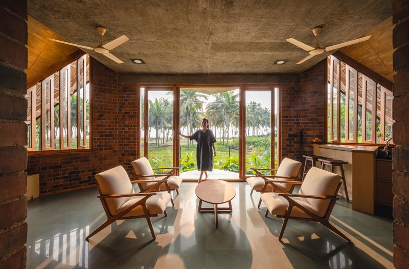 Living room with red brick walls and tall slatted windows framing a palm grove with a figure at the glass door