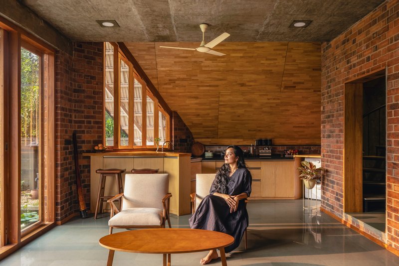 Kitchen area with angled timber ceiling panels and continuous slatted windows along the brick wall with a seated figure