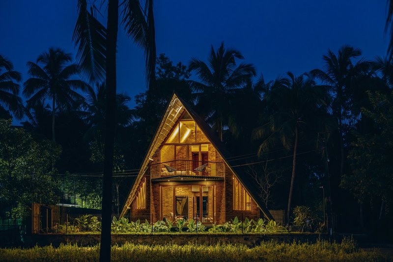 Night view of the steep triangular gable with timber cladding and illuminated upper window surrounded by palm trees
