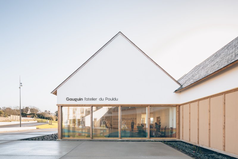 White gabled volume with full-height glazing revealing the interior workshop space beneath an overcast sky