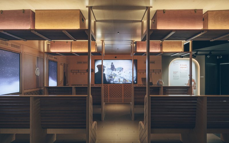 Exhibition gallery with timber-clad benches, overhead mezzanine structure and illuminated video screens at night