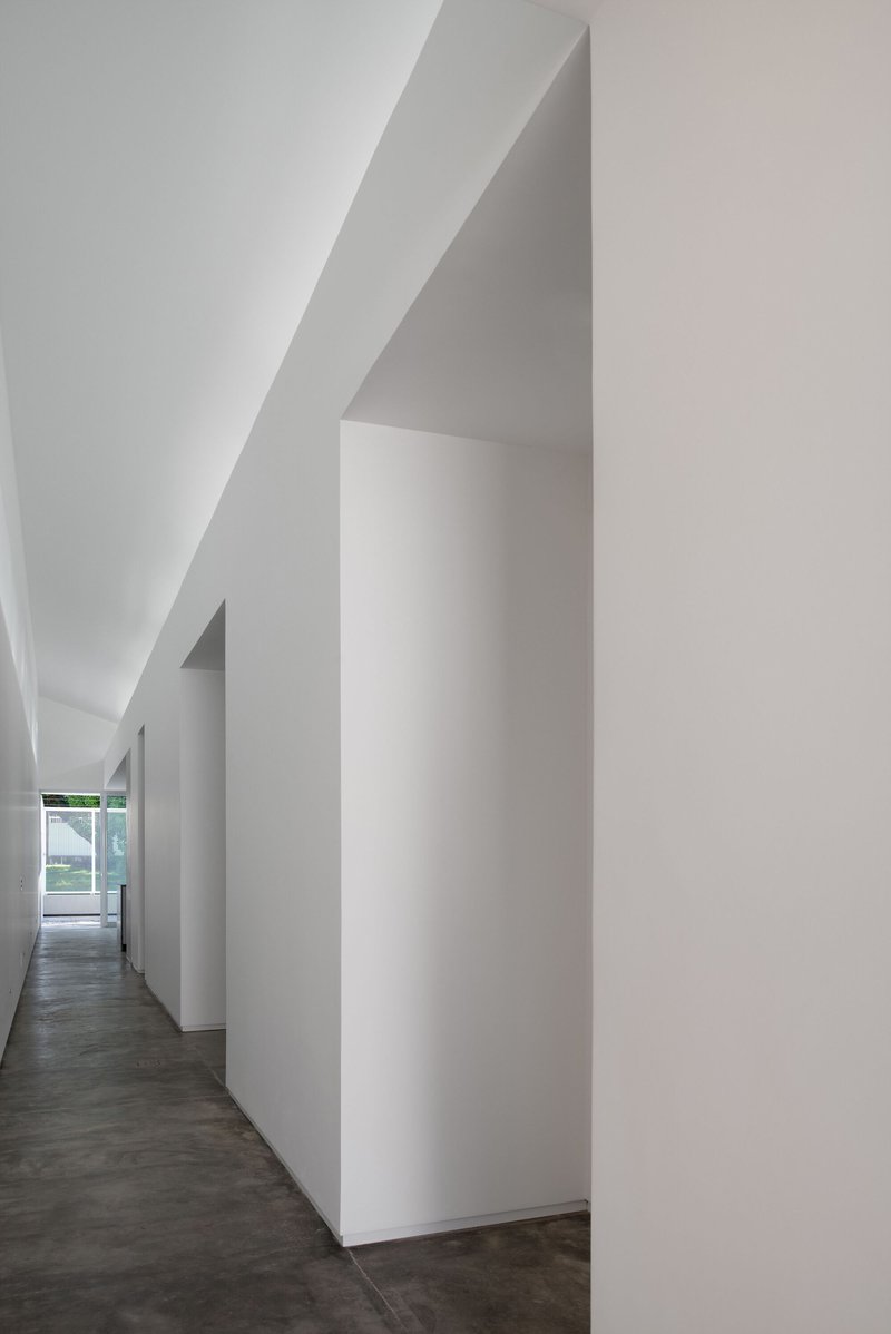 Narrow corridor with angular white walls and sloped ceiling leading toward a distant window