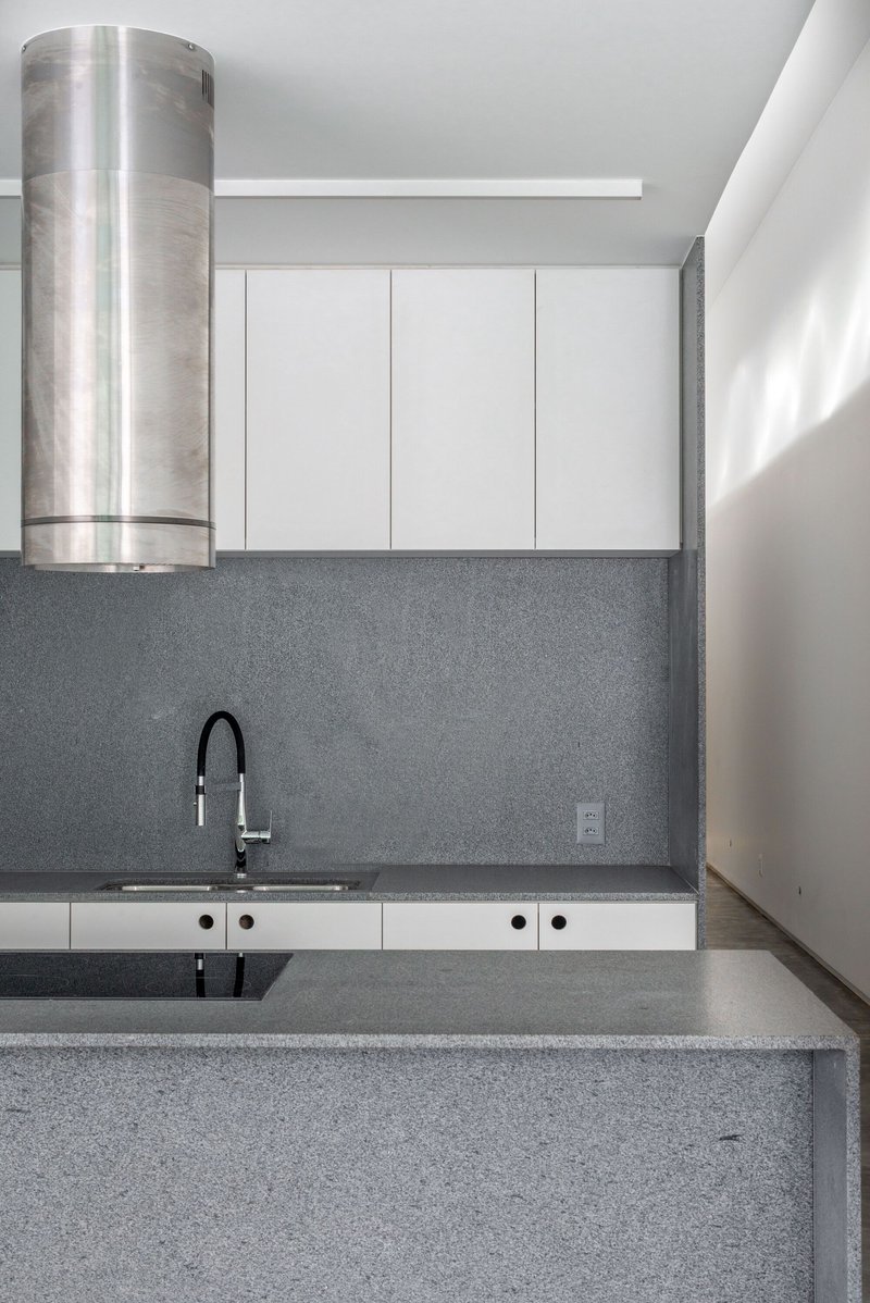 Kitchen with grey terrazzo countertops, white cabinets, cylindrical steel range hood, and sunlight entering from the right