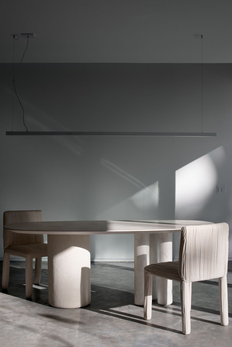 Dining area with circular table, ribbed chairs, and overhead linear pendant casting shadows on the wall