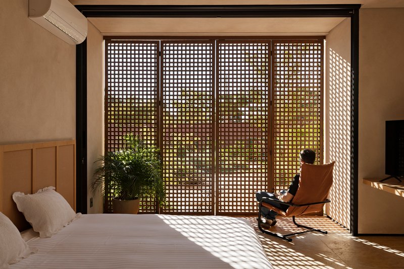 Bedroom with perforated wooden sliding doors filtering dappled sunlight onto a person seated in a lounge chair