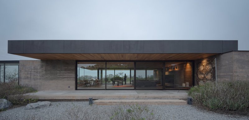 Front facade with recessed glazing beneath a cantilevered dark metal roof overhang and rammed earth walls