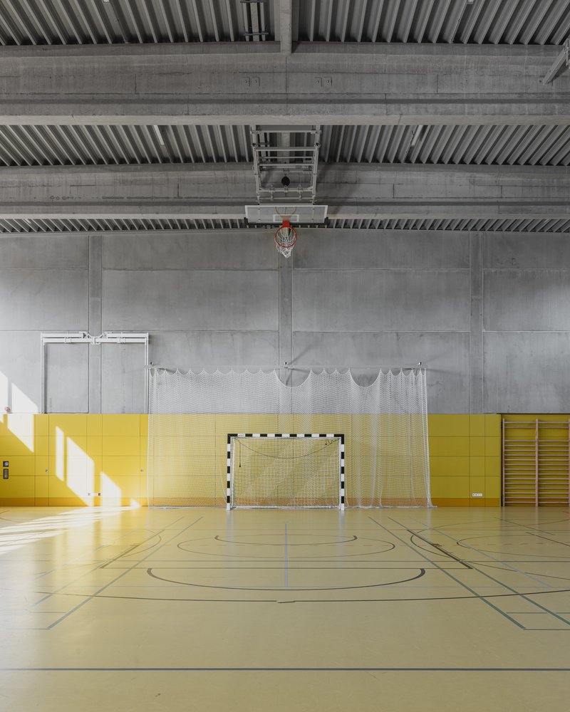 Gymnasium interior with basketball hoop, goal net, exposed concrete ceiling, and yellow wall panels with sunlight