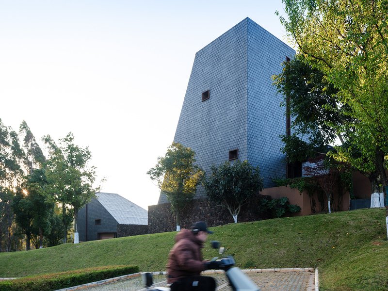 Shingled tower with punched windows overlooking sloped lawn as motorcyclist passes below at twilight