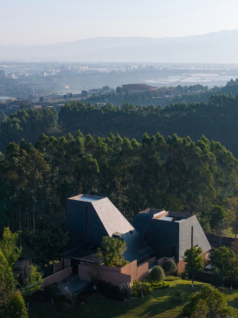 Aerial view of shingled volumes with terracotta base nestled in forested hillside above valley