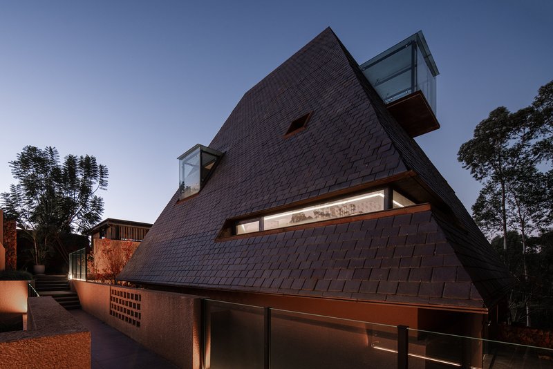 Steeply pitched shingle facade with horizontal window slits illuminated at twilight