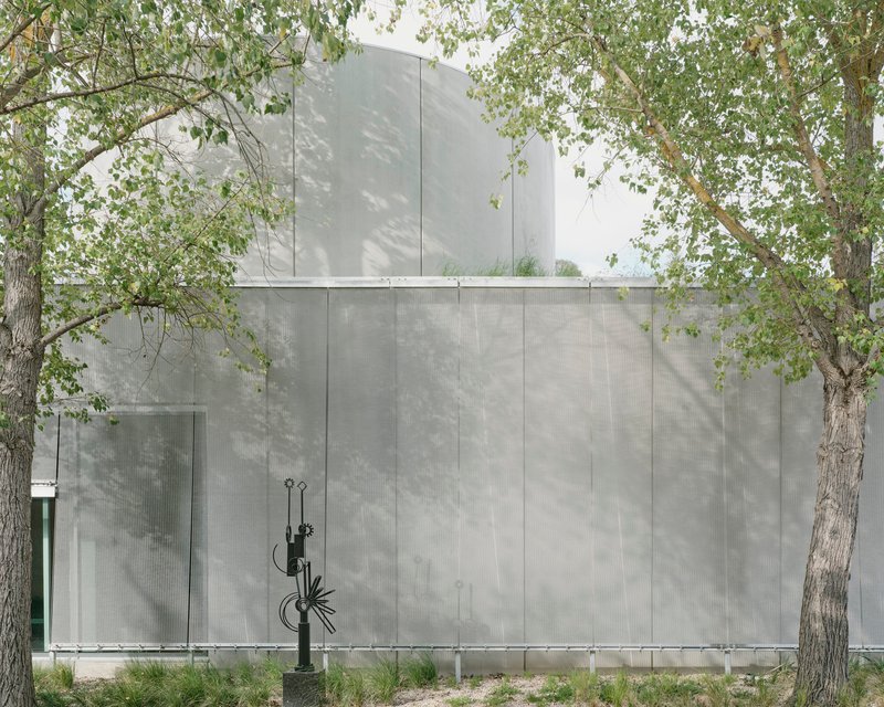 Textured concrete wall with vertical paneling beneath a protruding upper volume visible through dappled tree shadows