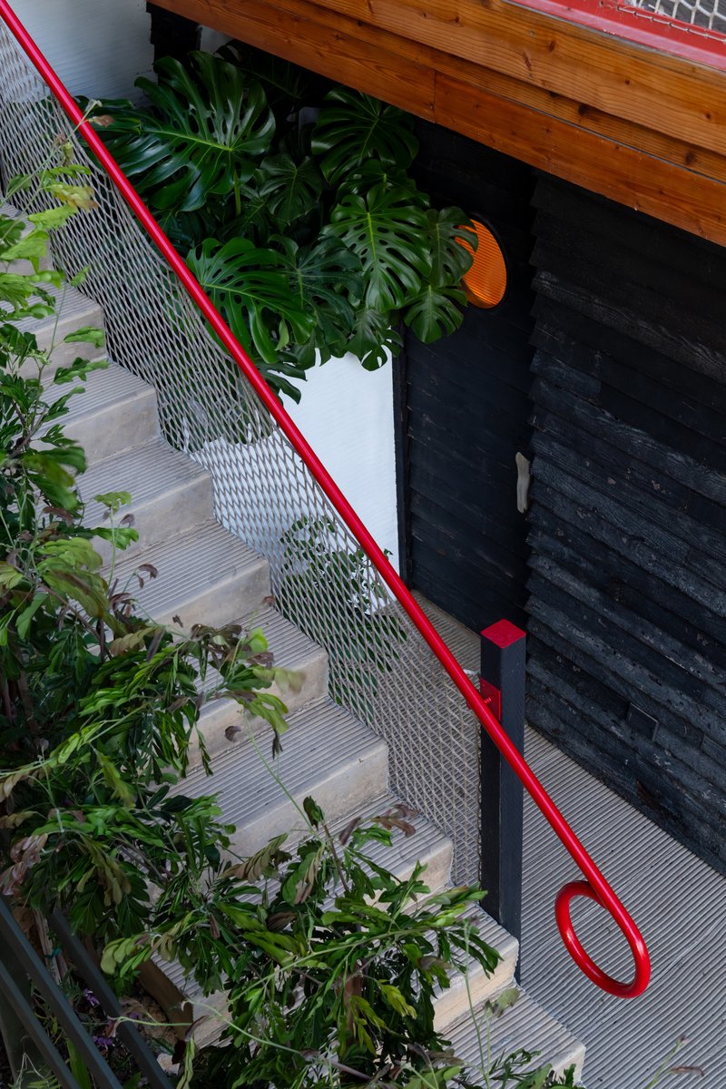 Staircase with red tubular handrail and monstera plants beside a charred timber wall