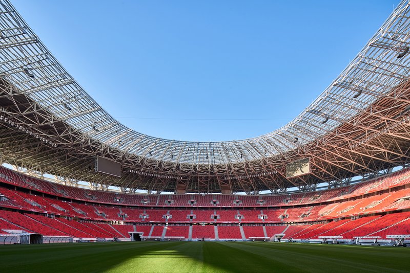 Curved steel truss roof structure spanning over red seating sections and the green striped playing field