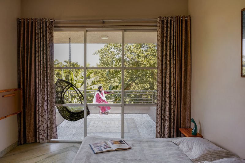 Bedroom view through sliding glass doors to a balcony where a person sits in sunlight