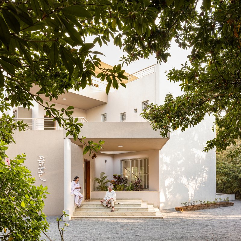 Front entrance steps with two figures seated beneath mature mango tree branches filtering afternoon light