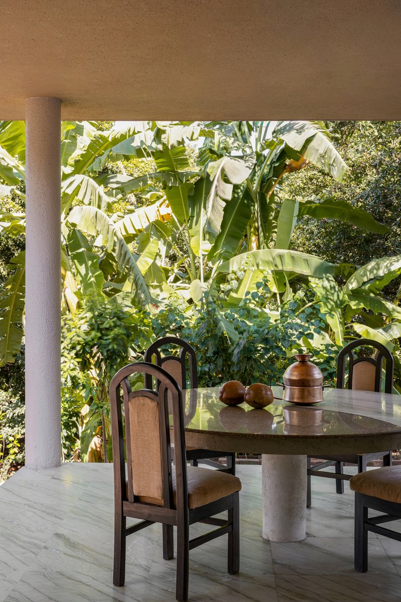 Covered terrace with cylindrical column and dark wood dining chairs facing dense banana plants in bright sunlight