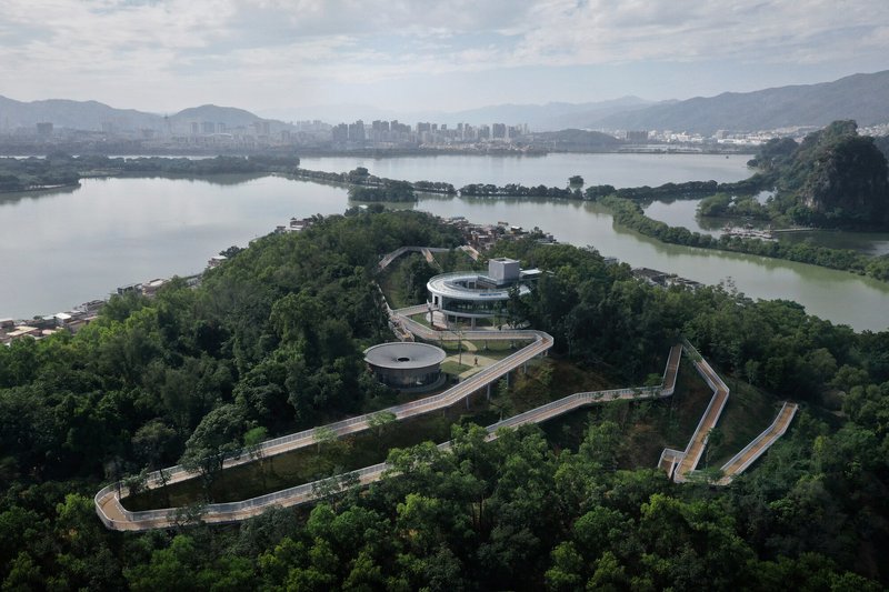 Aerial view of a zigzagging walkway connecting a circular pavilion on a forested peninsula surrounded by water