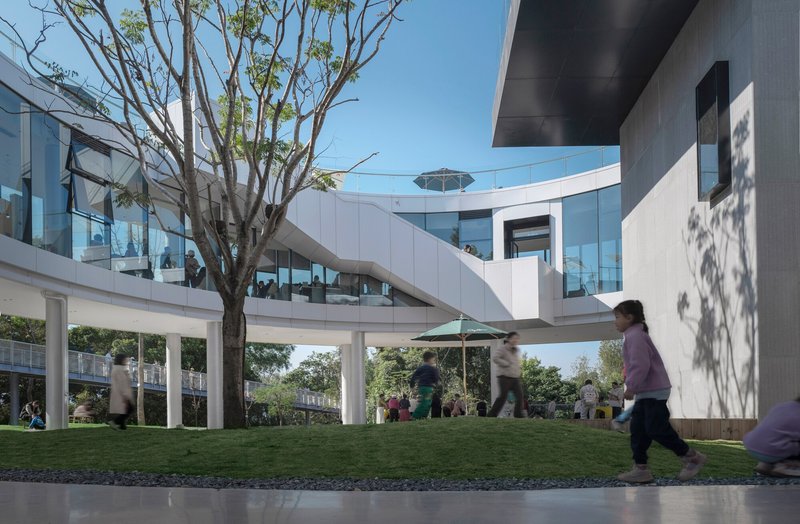 Sloped lawn with children running beneath curving white structures and a bare tree under clear sky