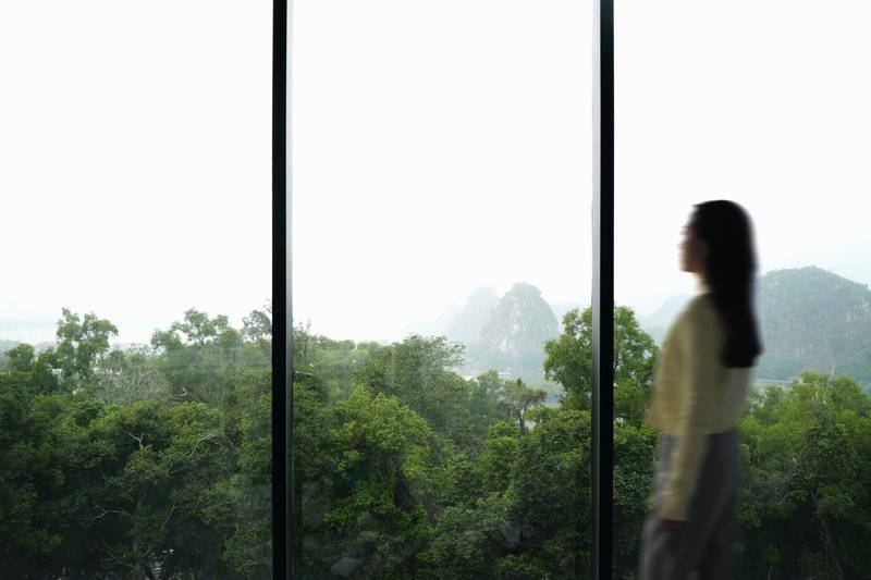Floor-to-ceiling window framing a forested valley with karst peaks visible through morning haze as a figure passes