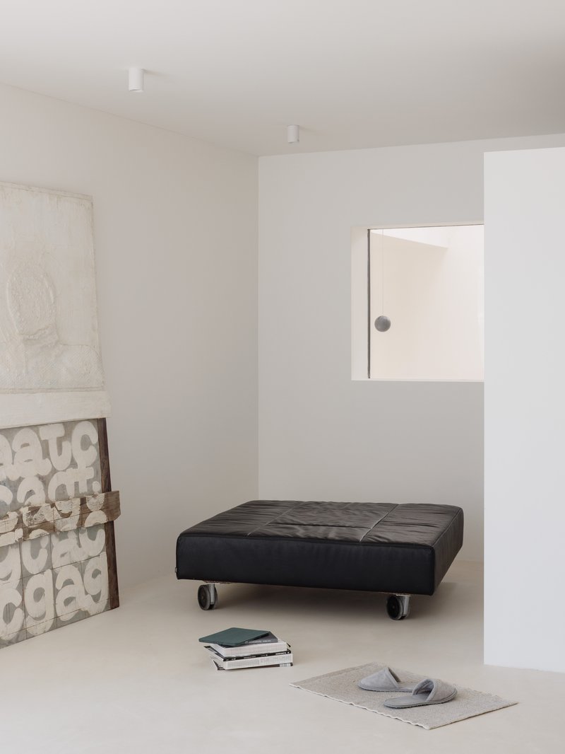 White interior room with a black tufted daybed on casters and stacked books on pale flooring