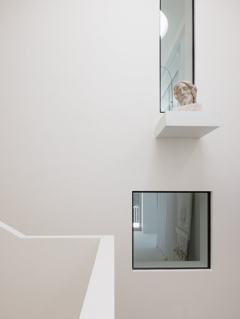 Sculptural bust on a floating shelf above a framed window opening in a white stairwell