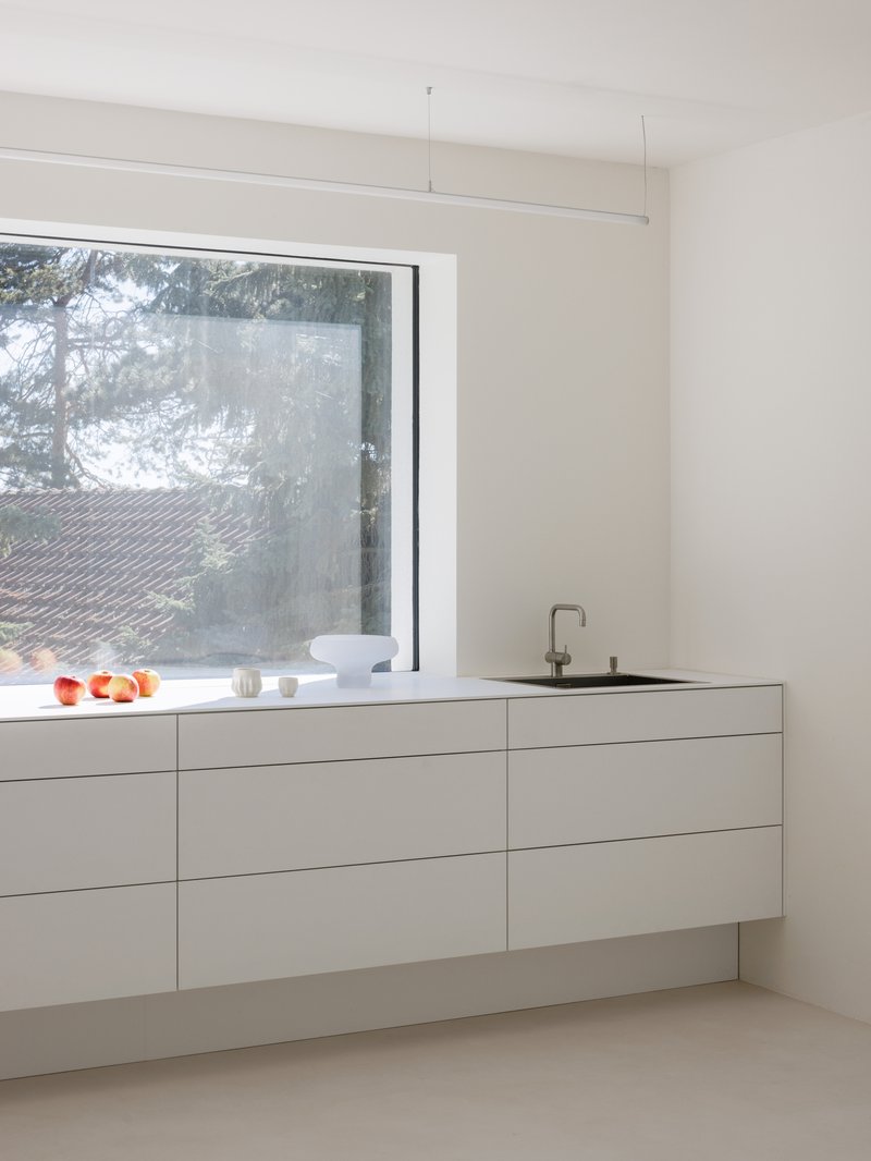 White handleless kitchen cabinetry beneath a horizontal window overlooking pine trees in soft light