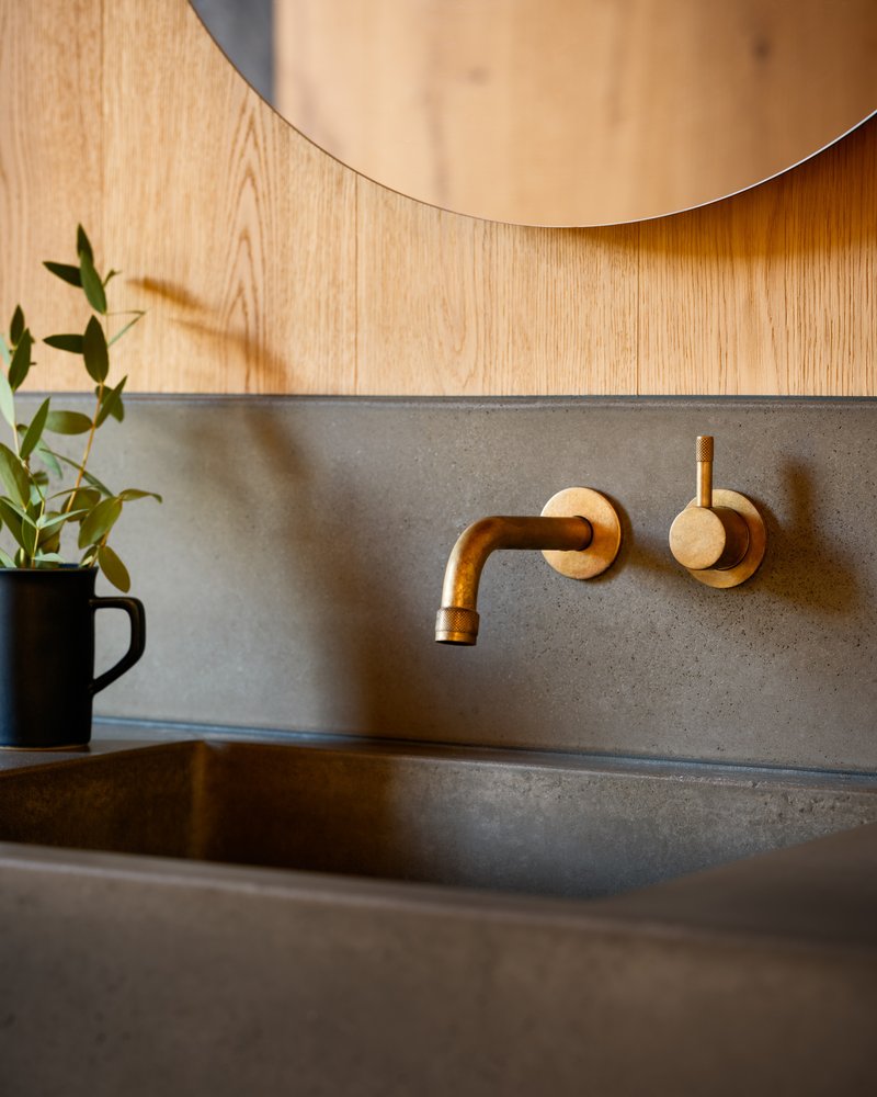 Close-up of brass wall-mounted faucet against concrete backsplash with black mug and greenery