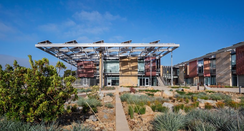Front facade showing corrugated metal panels and glass walls beneath solar canopy framed by native plantings