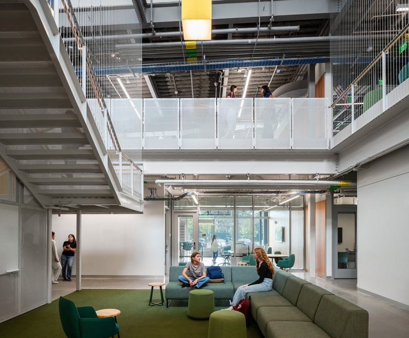Multi-level atrium showing steel staircase, bridge walkway, and lounge seating beneath exposed mechanical systems
