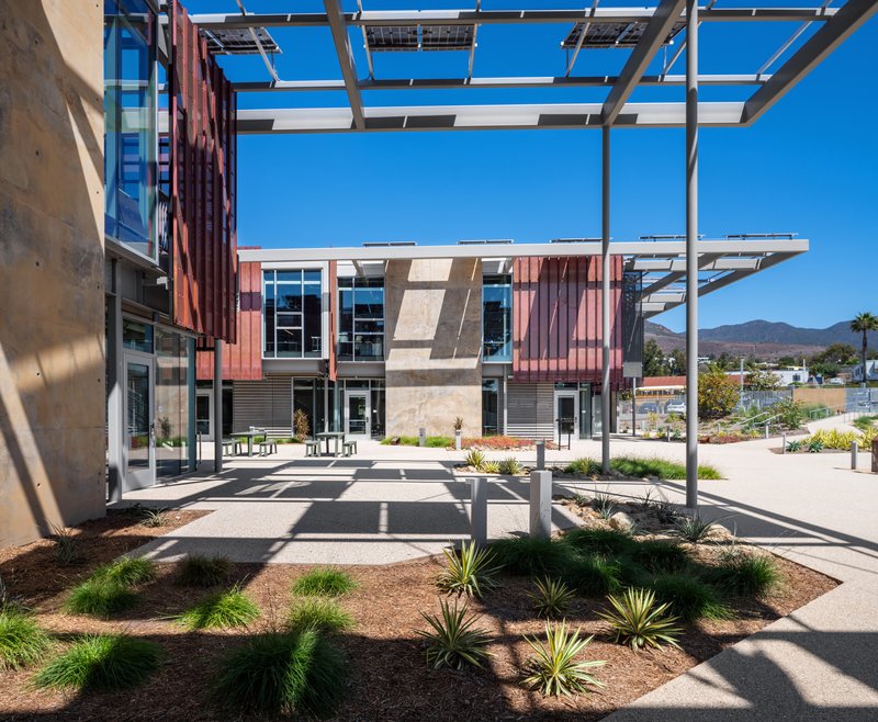 Central plaza with white steel pergola structure casting shadows over planted beds and paving