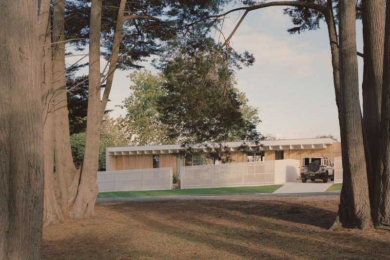 Long cream brick facade with exposed timber rafters viewed through a grove of mature trees at dusk