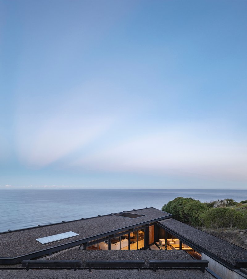 Angled gravel roof with skylights and glazed openings set into coastal vegetation at dusk