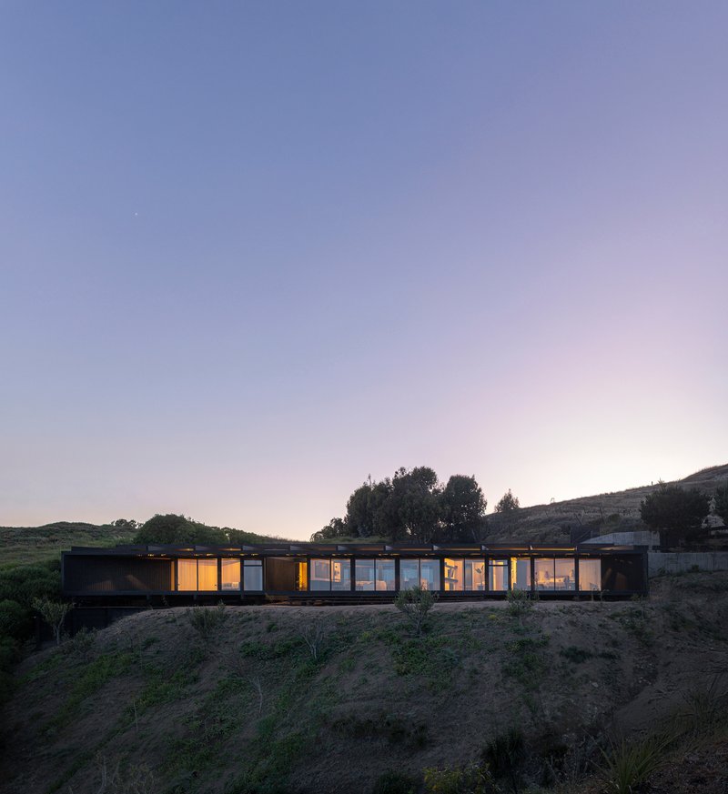 Illuminated linear volume set into a hillside viewed at dusk with rolling terrain and trees beyond
