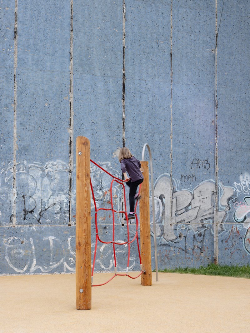 Child climbing a red rope net structure framed by timber posts against a blue tiled wall