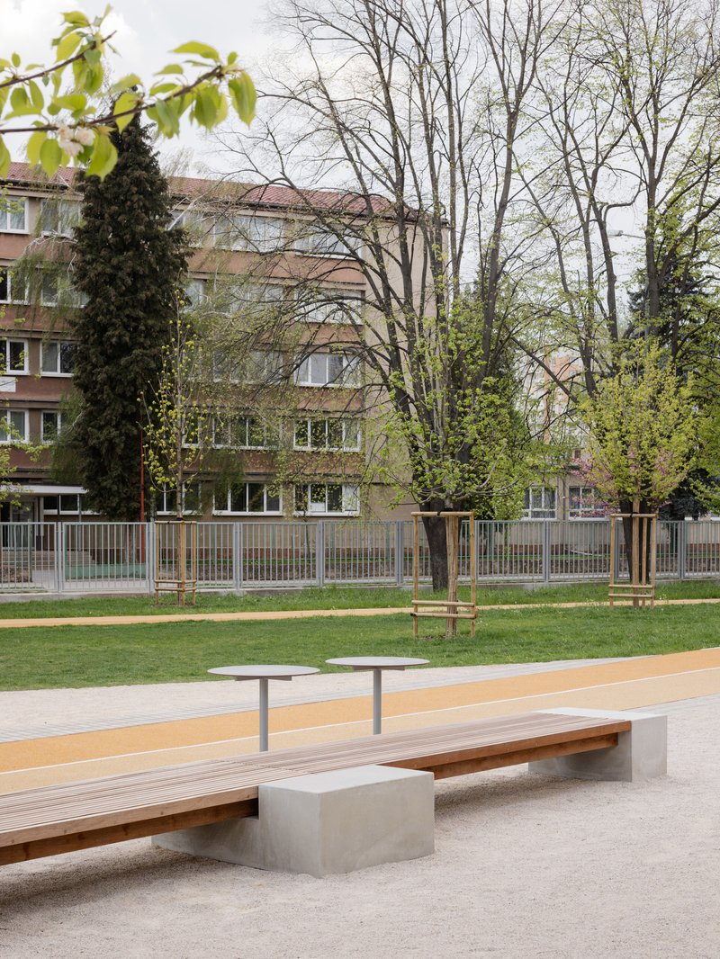 Timber and concrete bench beside a gravel play area with trees and school building in spring