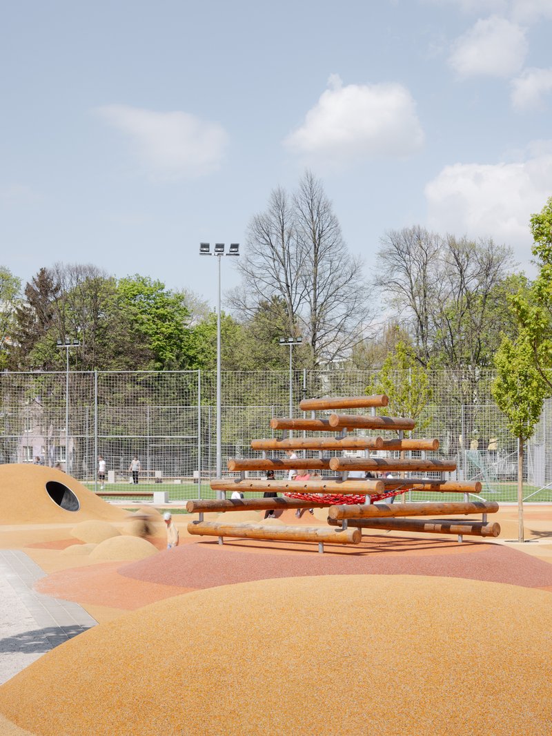 Timber play structure with stacked horizontal slats on rubberized orange surfacing beneath bare trees