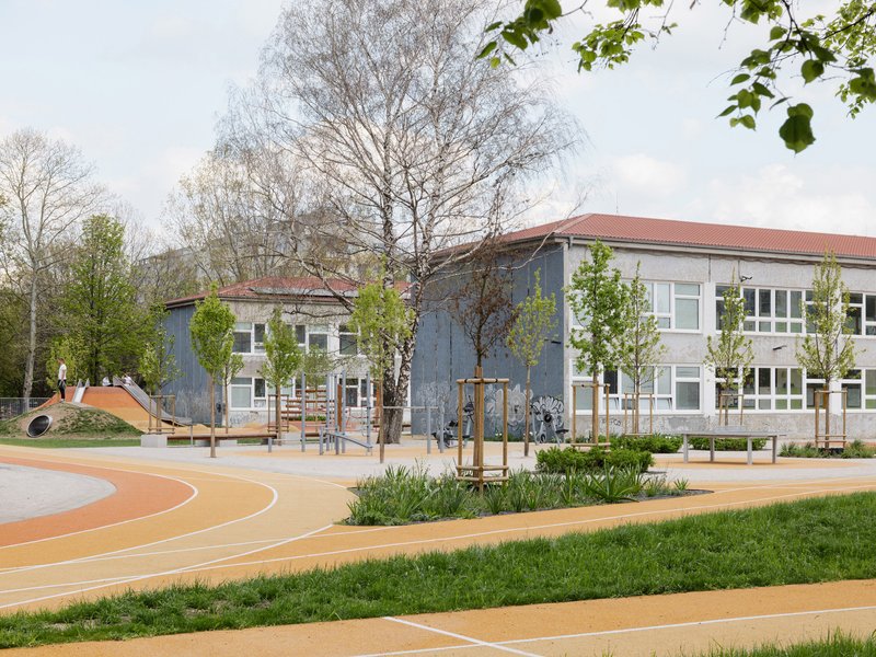 School courtyard with orange play surfaces, planted beds, and young trees beneath an overcast spring sky