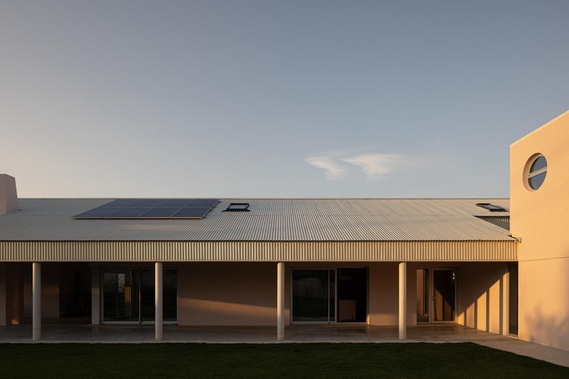 Long facade with corrugated metal roof, solar panels, and colonnade overlooking a gravel courtyard