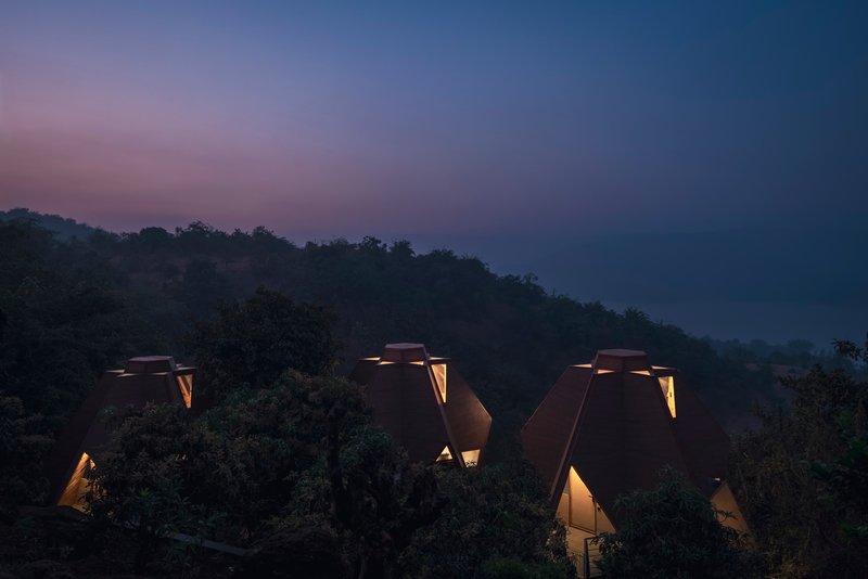 Group of triangular roof forms with glowing apertures nestled among trees at twilight