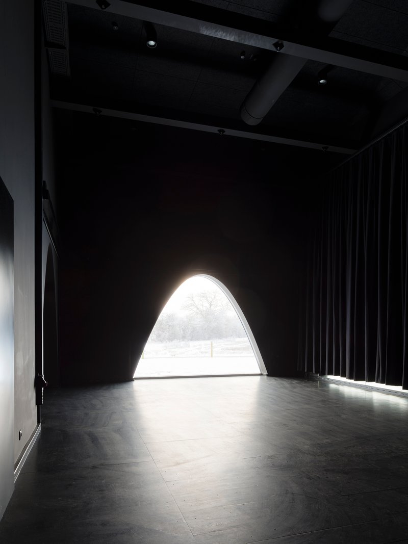 Arched opening flooding light into a dark performance space with black curtains and exposed ceiling