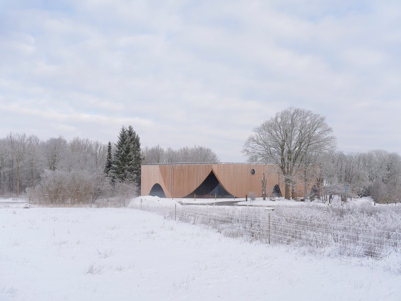 Wood-clad volume with arched openings set in a snow-covered field beneath bare winter trees