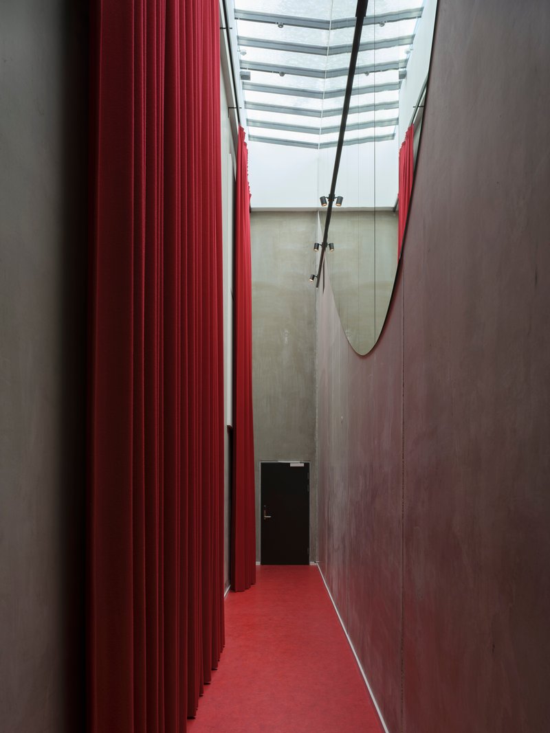 Narrow corridor with red ribbed wall and matching floor beneath a skylight with suspended trapeze
