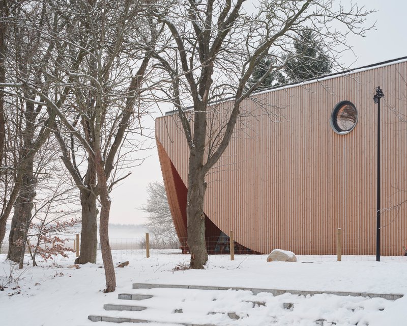 Vertical timber facade with circular window and diagonal entrance beneath bare winter trees